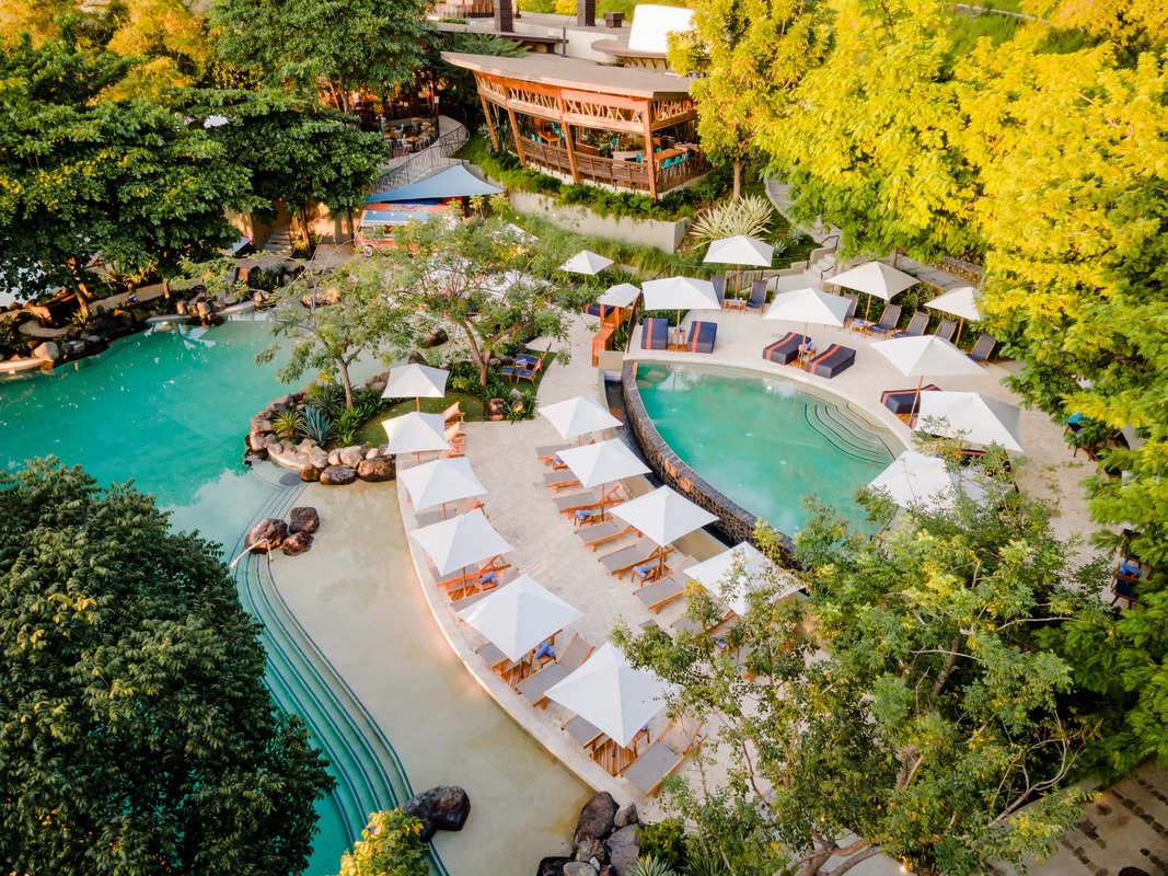 looking down at two pools surrounded by trees and cabanas 